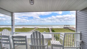 A covered outdoor seating area with white chairs and a table, overlooking a beach with a boardwalk and a pier.