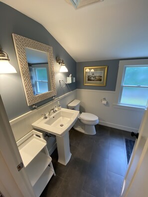 Une salle de bain avec un lavabo, des toilettes et un meuble-lavabo, tous de couleur blanche. On y trouve un miroir avec un cadre décoratif, un porte-serviettes et un tableau encadré accroché au mur.