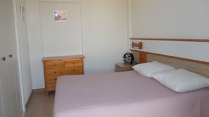 A bedroom with a bed, wooden dresser, bedside table with a fan, and a door with a picture.