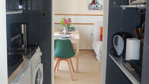 A compact kitchen with a washing machine, a dining area with a green chair, and a dining table with a vase of flowers.
