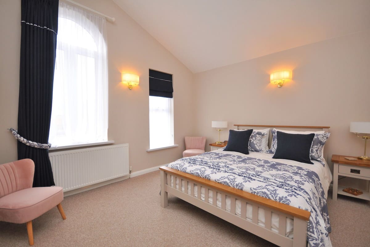 Een slaapkamer met een bed, twee bedlampen, een raam met gordijnen, een radiator en een roze stoel.