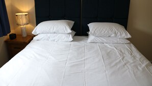 A double bed with white bedding and a dark headboard, a bedside table with a lamp, and a wooden nightstand.