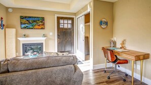 A living room with a fireplace, a sofa, a desk with a chair, and a painting on the wall. - Shore 2 Please - Lower Unit,BRIGHT/Mountain views (Port Renfrew)
