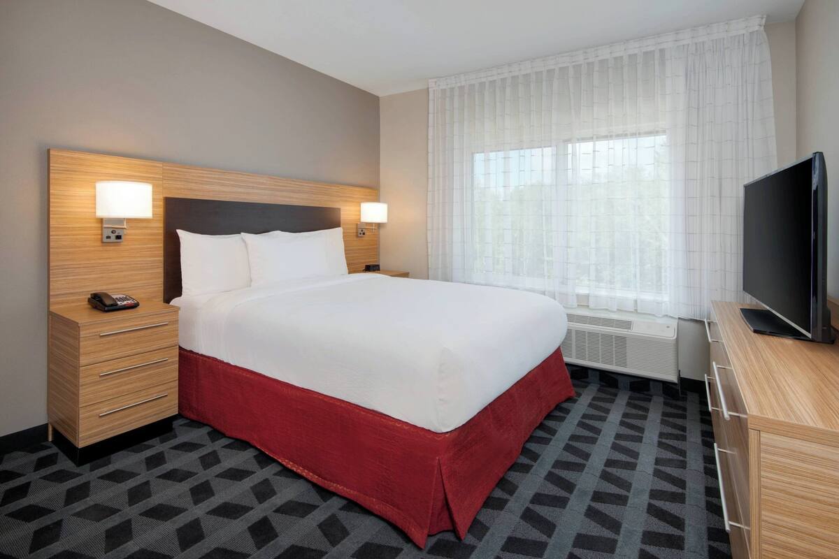 A hotel room with a large bed, a TV, a desk, and a window with curtains.