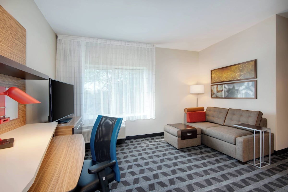 A hotel room with a desk, chair, sofa, TV, lamp, and artwork on the wall.