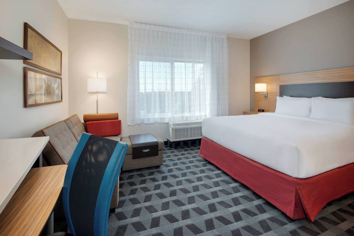 A hotel room with a bed, a desk, a chair, a small table, a lamp, and a window with sheer curtains.