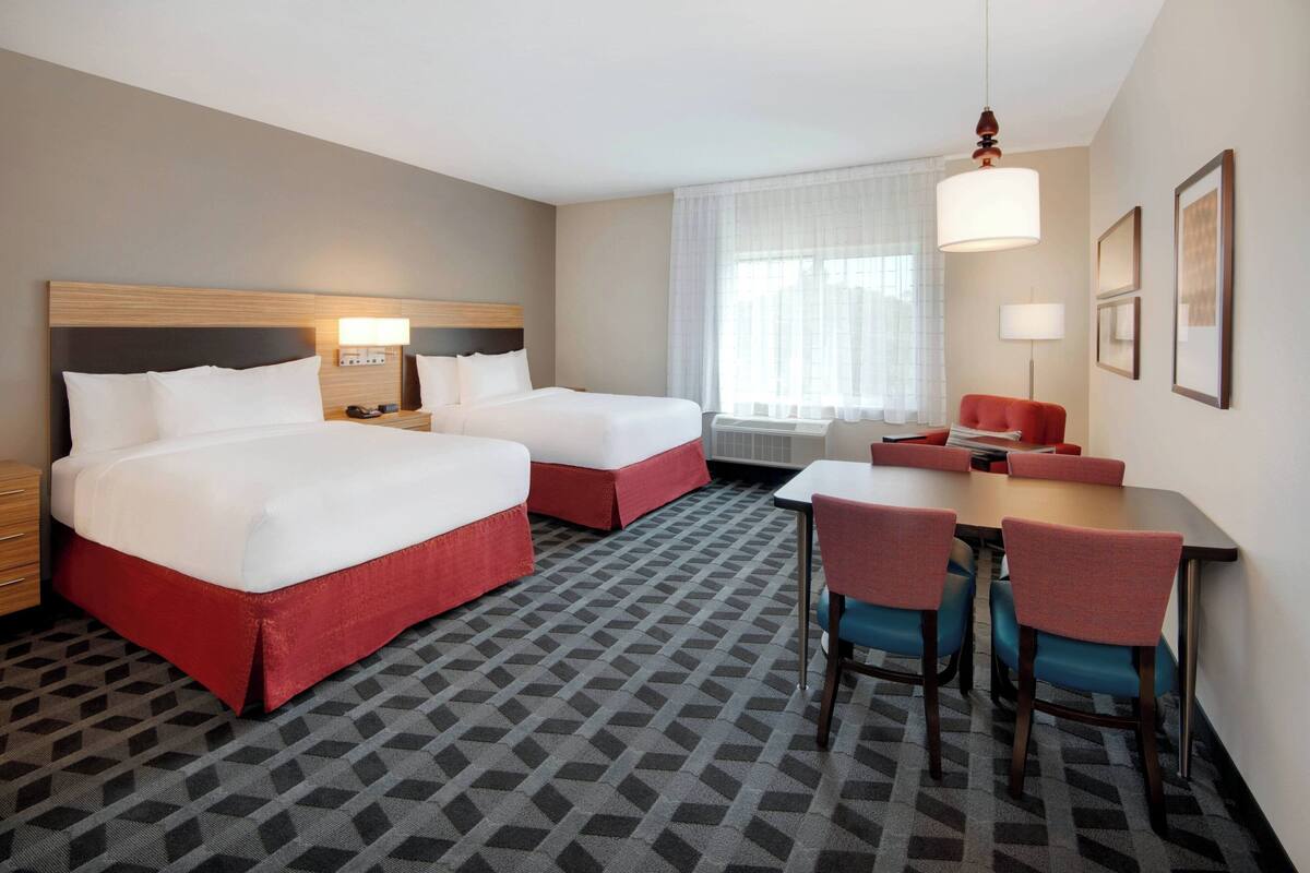 A hotel room with two beds, a dining table with chairs, a desk, and a large window with curtains.