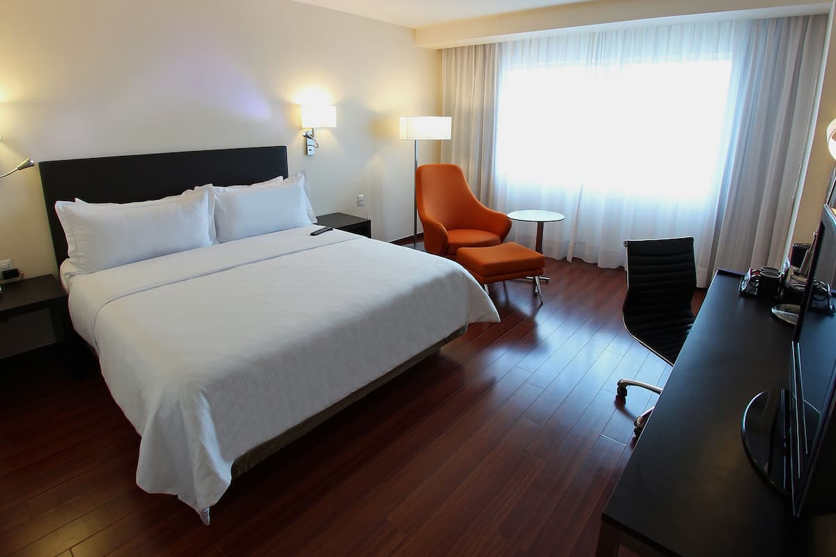 A hotel room with a large bed, a desk with a chair, a lamp, and a view of the outdoors through a window with curtains.
