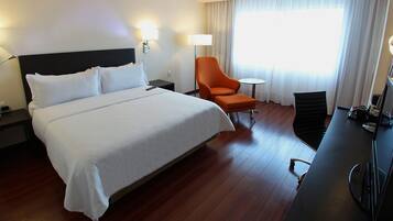 A hotel room with a large bed, a desk with a chair, a lamp, and a view of the outdoors through a window with curtains.