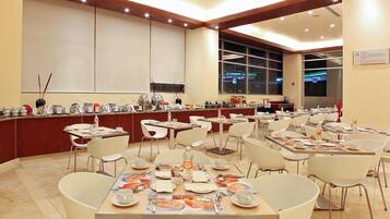 A spacious dining area with multiple tables and chairs, a buffet counter with various food items, and a large window providing a view of the cityscape.