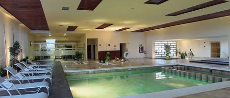 A modern indoor swimming pool with lounge chairs, wooden ceiling beams, and potted plants.