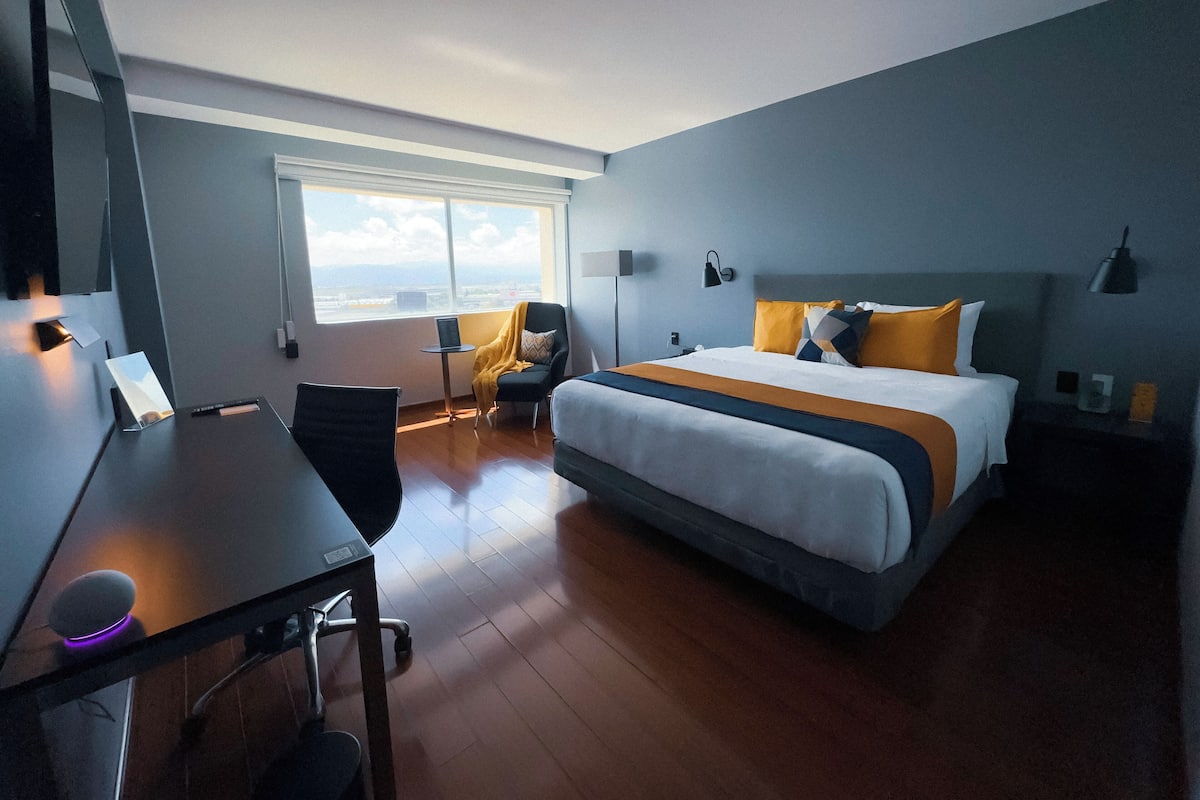 A modern hotel room with a large bed, a desk, a chair, and a window offering a view of the city.
