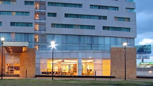 Un hôtel à plusieurs étages doté d’un design moderne, caractérisé par une grande façade vitrée, une entrée centrale avec un espace abrité et un escalier apparent menant aux étages supérieurs.