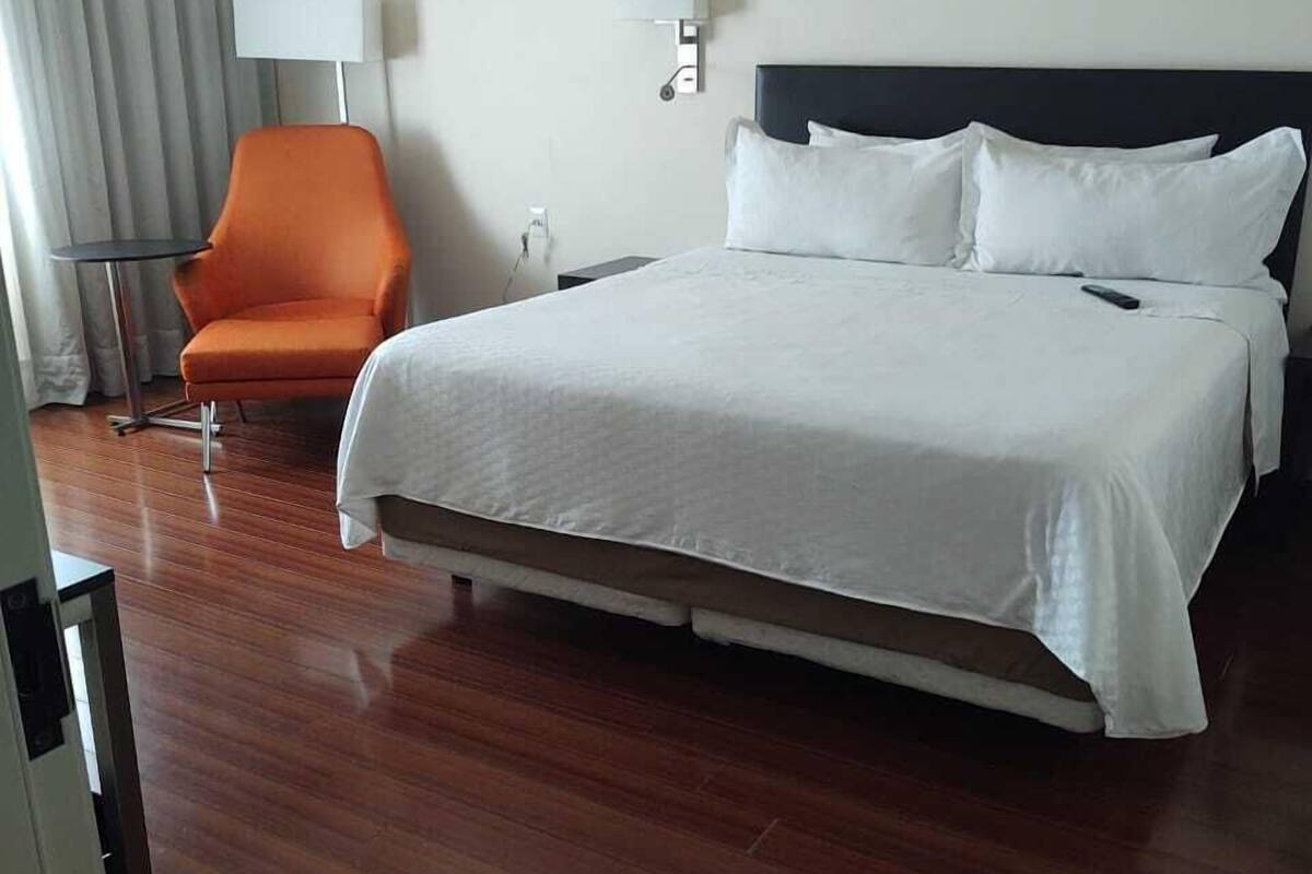 A hotel room with a large bed, an orange chair, a bedside table, and a lamp.