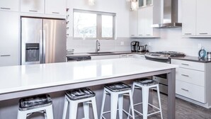 Una cocina moderna con armarios blancos, un refrigerador de acero inoxidable, una isla grande con taburetes, luces colgantes y un ventanal que proporciona luz natural.