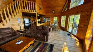 A cabin interior with a wooden staircase, a living room with a sofa and coffee table, a kitchen area, and large windows offering a view of the outdoors. - Best view in Georgia. Unbelievably peaceful. Amazing night sky. (Morganton)