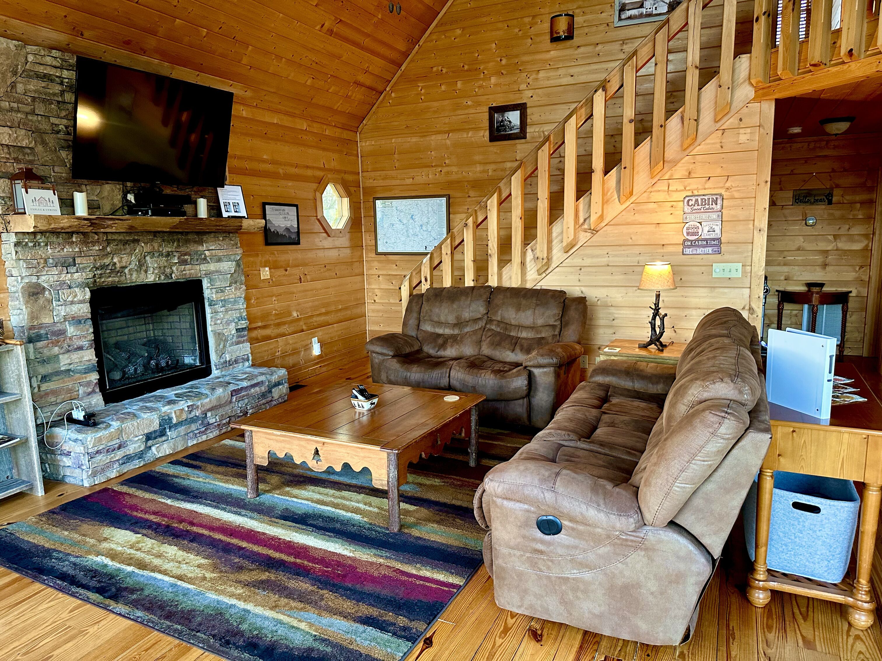 A cozy cabin interior with a stone fireplace, a large flat-screen TV, a comfortable sofa, and a wooden staircase.