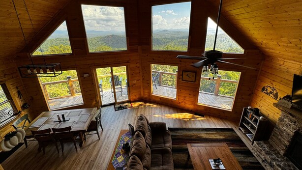 A spacious loft-style room with a high ceiling, large windows offering mountain views, a cozy seating area with a sofa and coffee table, and a fireplace. - Best view in Georgia. Unbelievably peaceful. Amazing night sky. (Morganton)