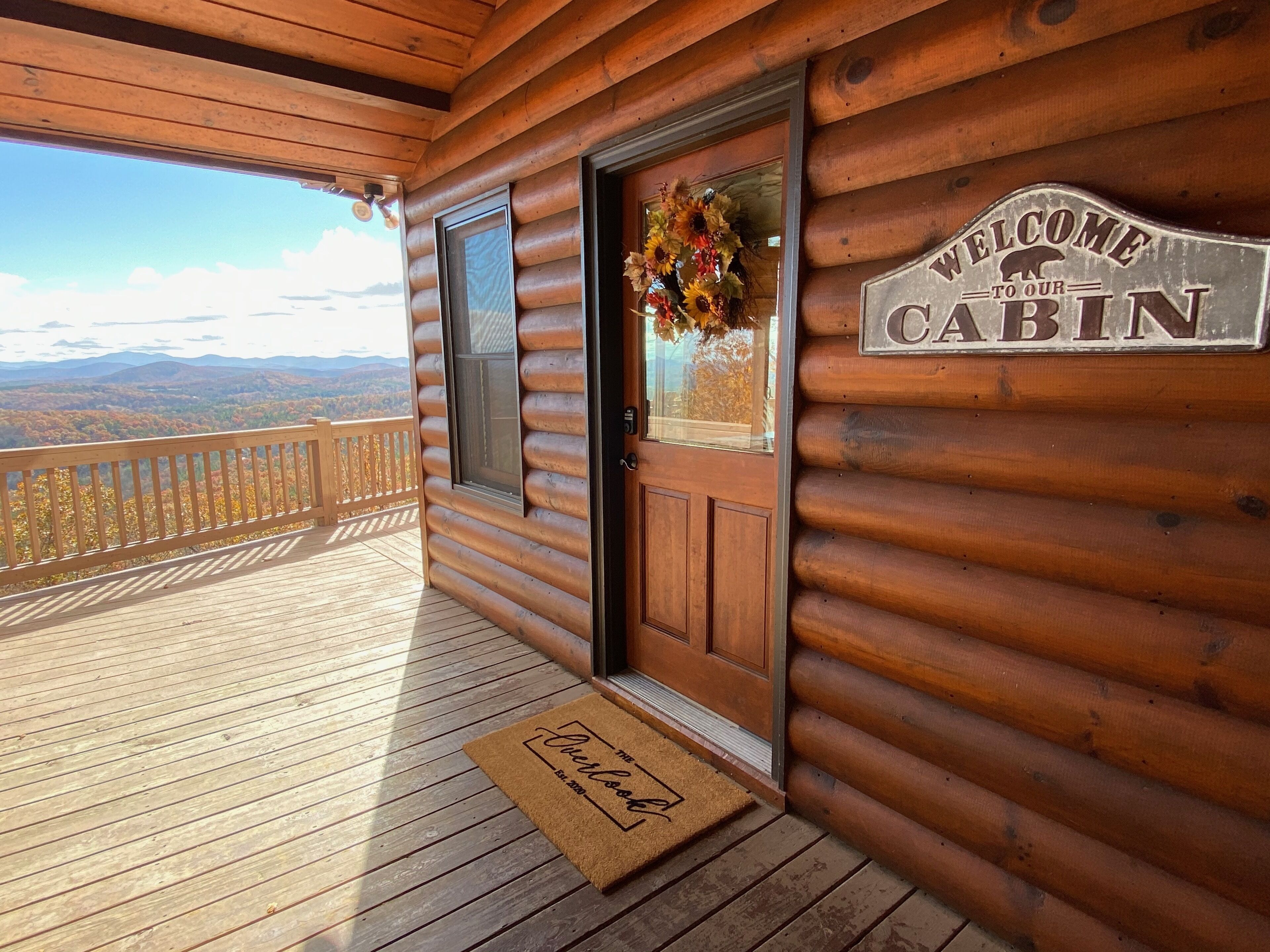 A wooden cabin with a welcome sign, a door with a wreath, and a view of the mountains.