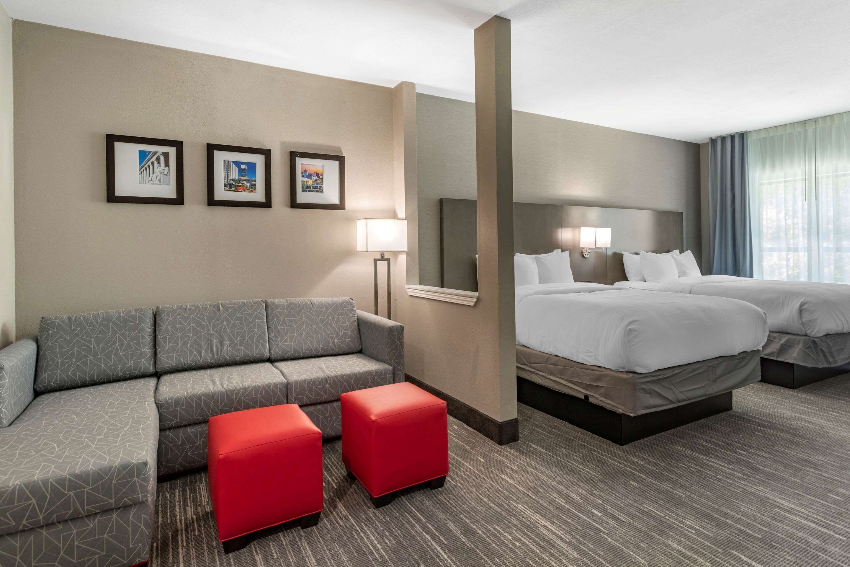 A hotel room with a grey sectional sofa, two red ottomans, a large bed with white bedding, and three framed pictures on the wall.