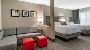 A hotel room with a grey sectional sofa, two red ottomans, a large bed with white bedding, and three framed pictures on the wall.