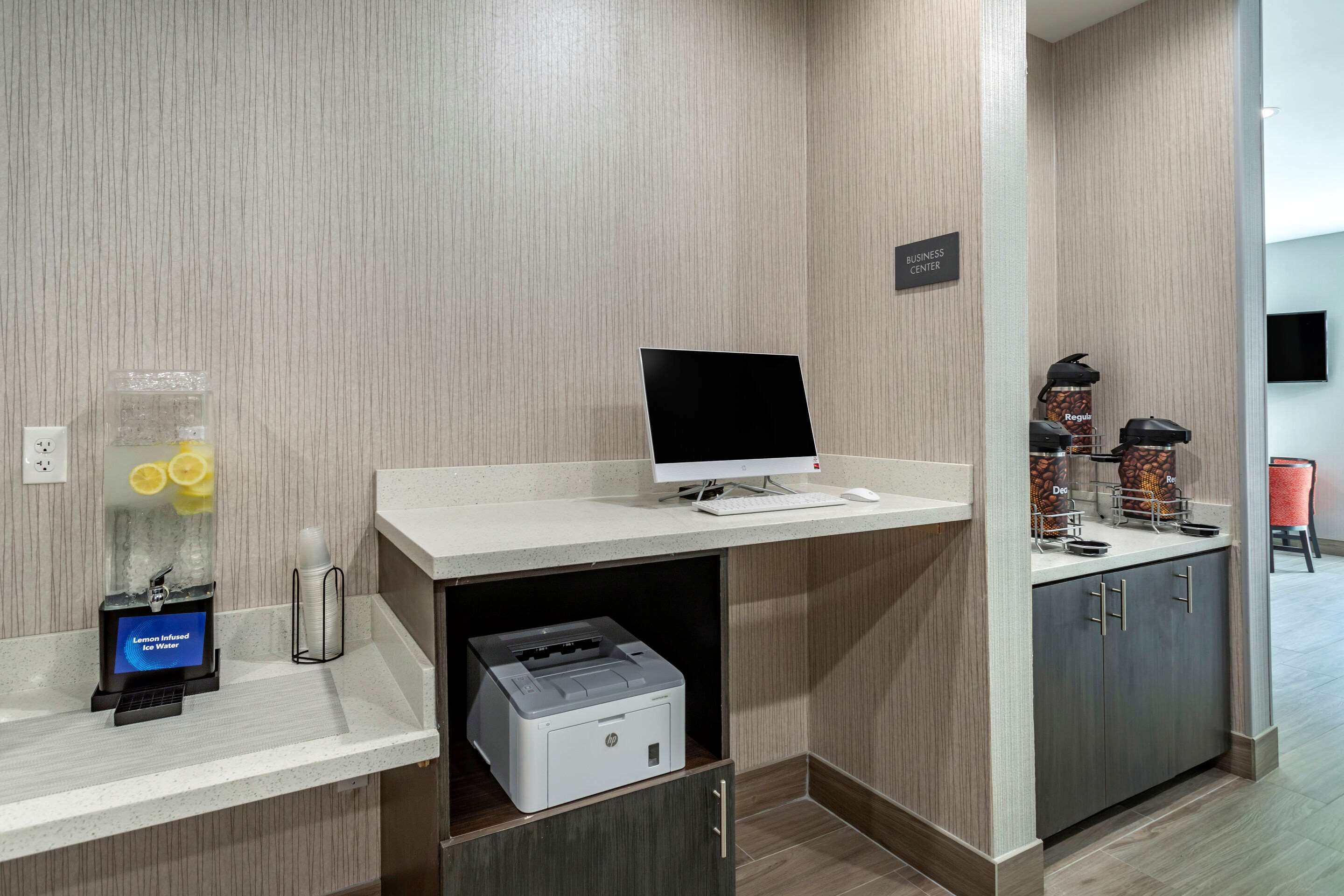 A business center with a desktop computer, printer, and beverage dispensers.