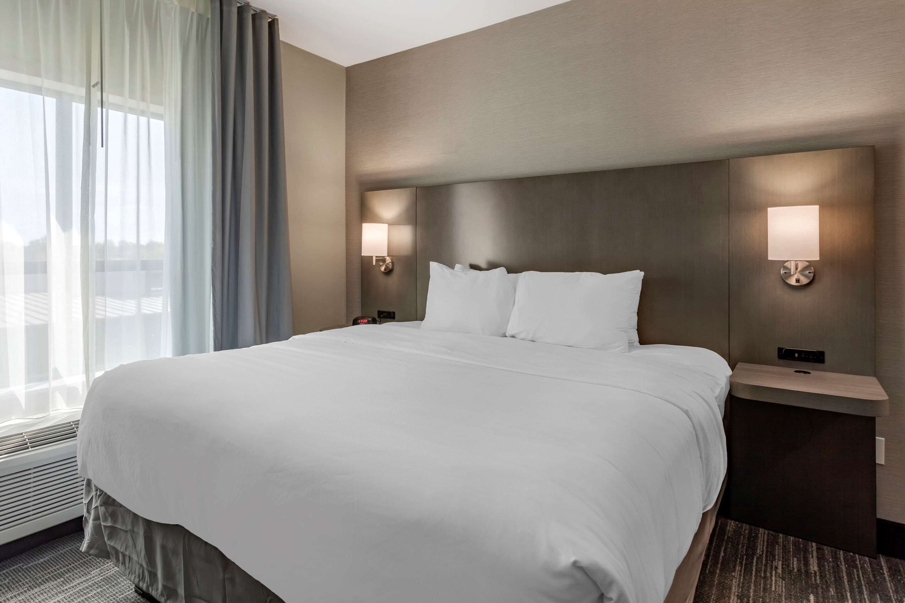 Una habitación de hotel con una cama grande, dos mesitas de noche con lámparas, una ventana con cortinas y una lámpara de pared.
