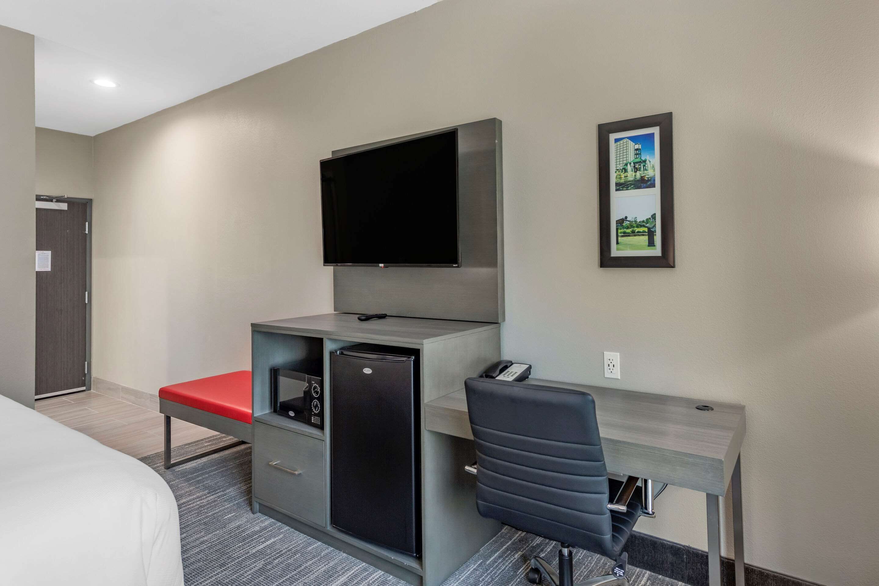 Habitación de hotel con cama, escritorio con silla, televisor montado en la pared, refrigerador y un cuadro enmarcado.