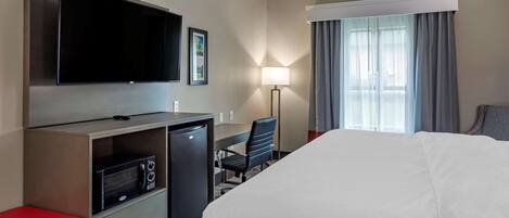 A hotel room with a bed, a desk with a computer, a TV, a microwave, a red bench, and a window with curtains.