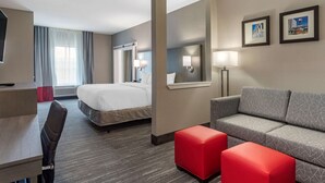 A hotel room with a bed, a desk, a sofa, and a red ottoman. There are two framed pictures on the wall. - Comfort Inn & Suites Olive Branch Old Towne (Olive Branch)