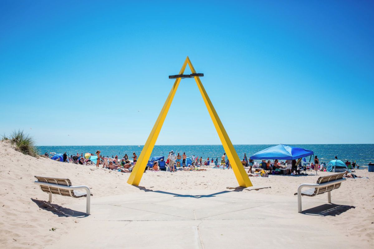 Une plage avec une structure triangulaire jaune, deux bancs et des personnes à l’arrière-plan.