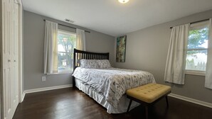 Quarto com uma cama, um banco e duas janelas com cortinas.