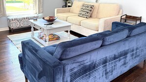 Uma sala de estar com um sofá bege, um sofá azul, uma mesa de centro e uma janela com cortinas.