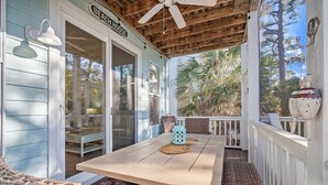 Un porche cubierto con una mesa y sillas de mimbre, un cartel que dice "Beach House" y vistas a palmeras.