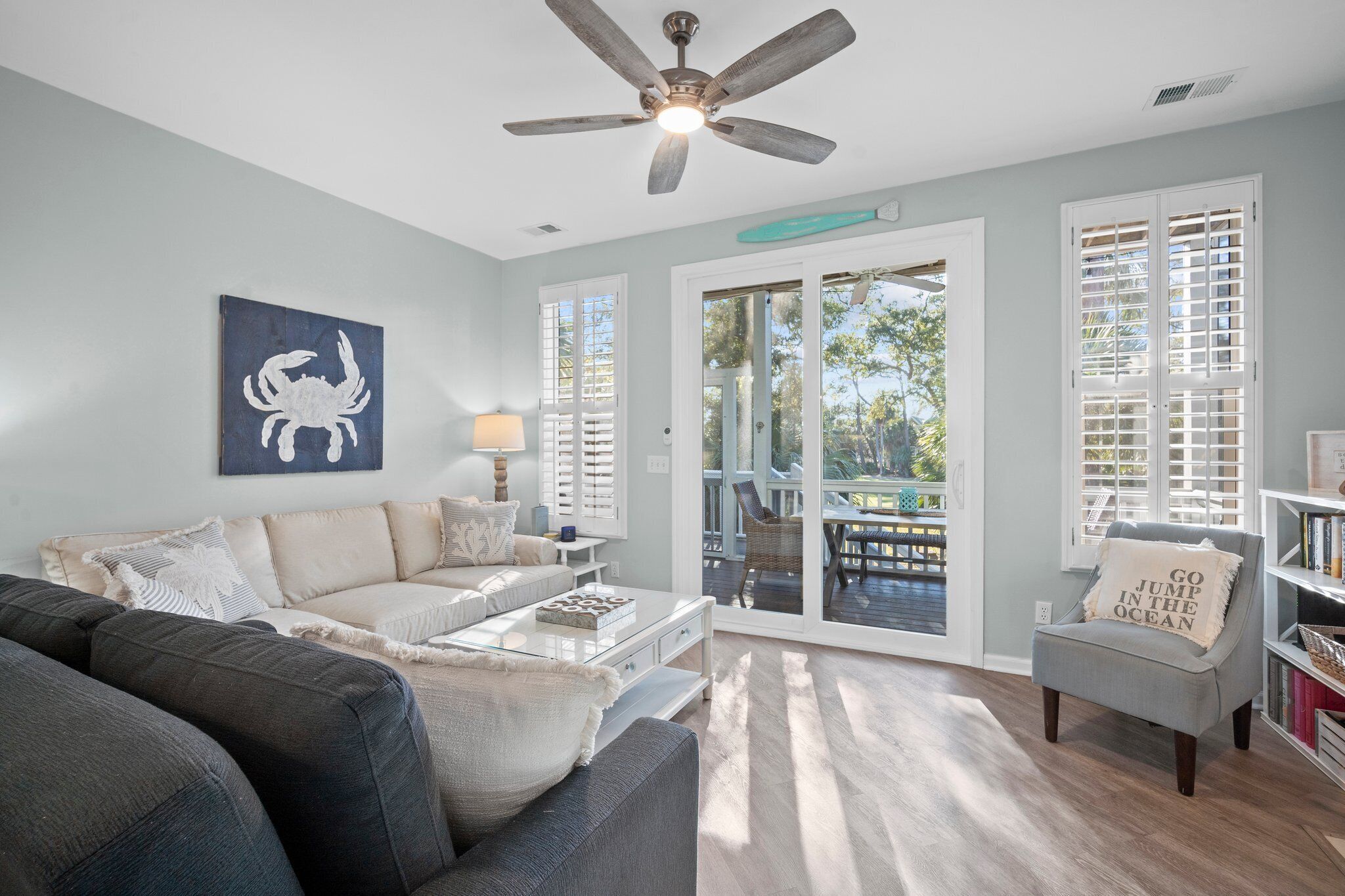 A living room with a sofa, armchair, and coffee table, a ceiling fan, and a view through glass doors to an outdoor deck.