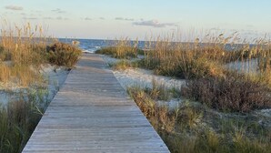 Una pasarela de madera que conduce a la playa, rodeada de arena y dunas cubiertas de hierba.