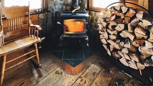 A cozy cabin interior with a wood-burning stove, a stone wall, and a stack of firewood. - Private Log Cabin Rental (Drayton Valley)