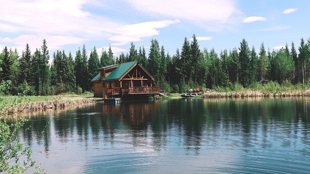 A cabin with a green roof, situated on a lake surrounded by dense forest. - Private Log Cabin Rental (Drayton Valley)