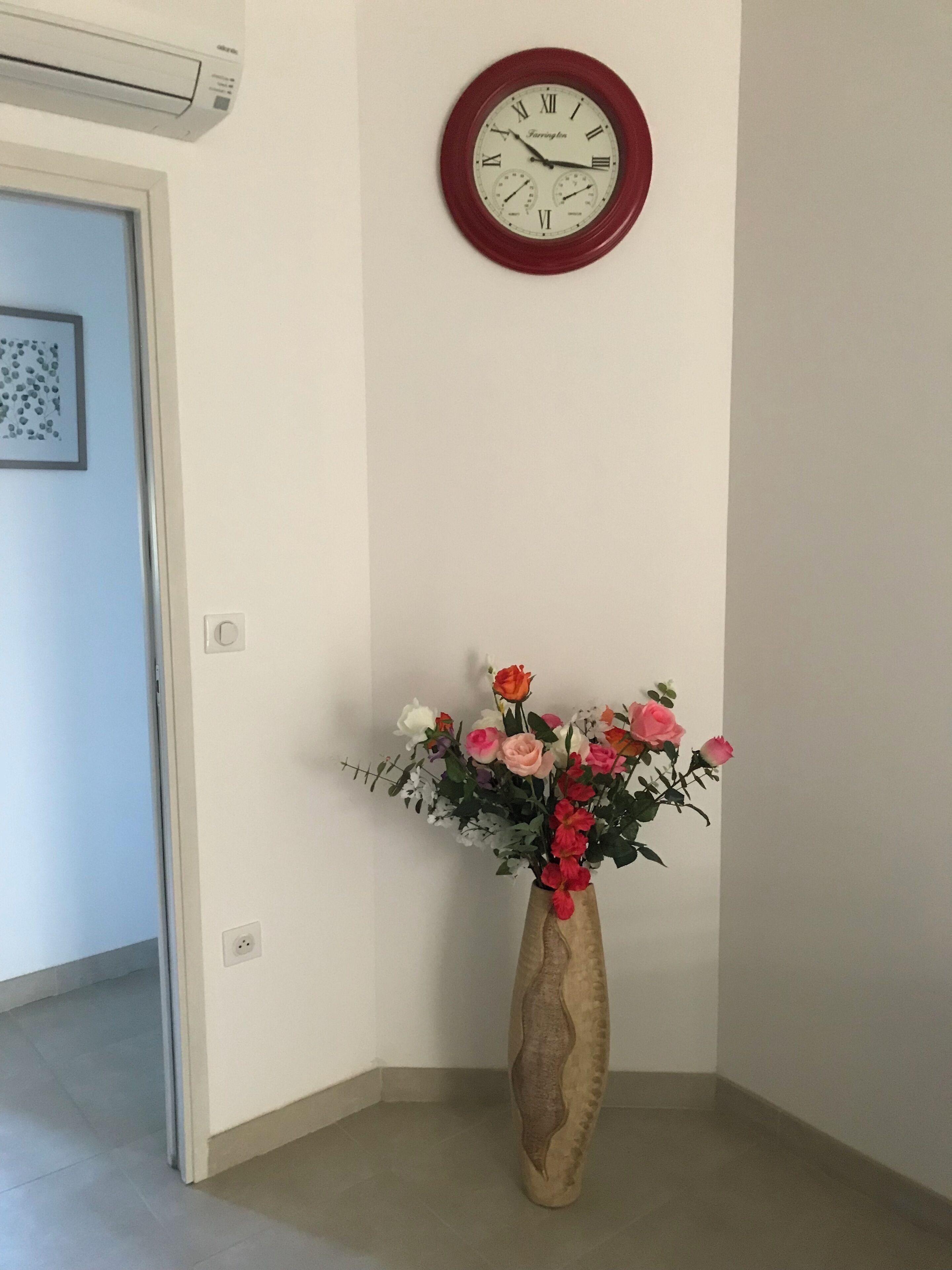 A hallway with a wall-mounted clock, a vase with flowers, and a door.
