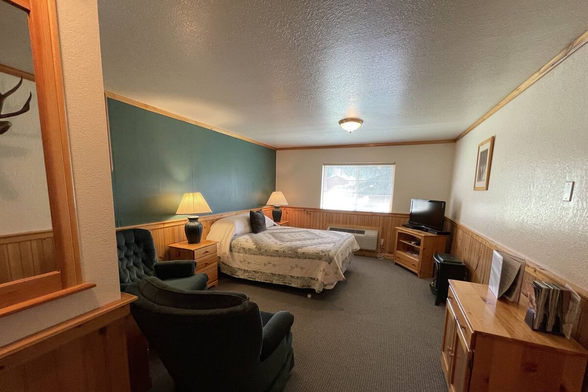 A small, cozy room with a bed, two bedside tables with lamps, a chair, a television, and a window with blinds.