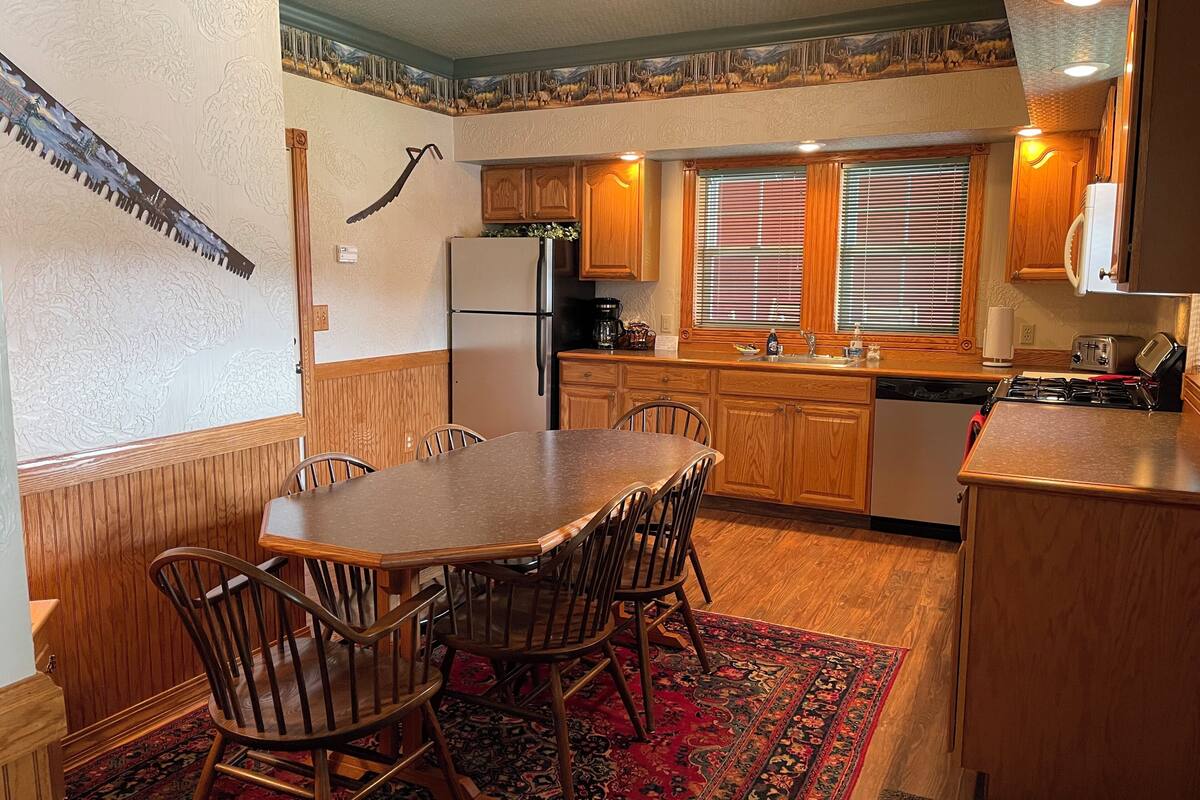 A kitchen with wooden cabinets, a dining table with chairs, a refrigerator, a stove, and a window with blinds.