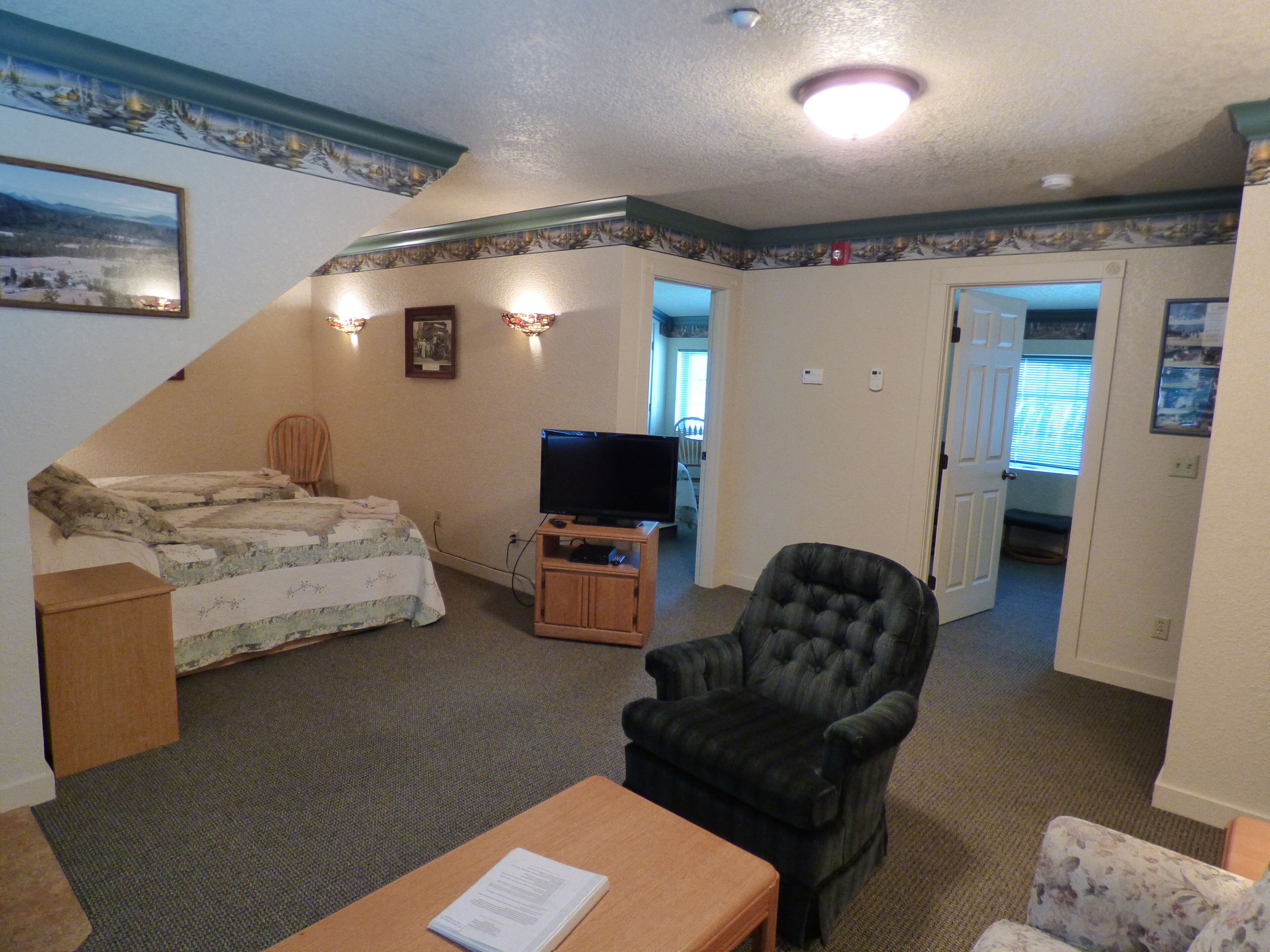 A bedroom with a bed, a television, a chair, and a desk. There is a door leading to another room.
