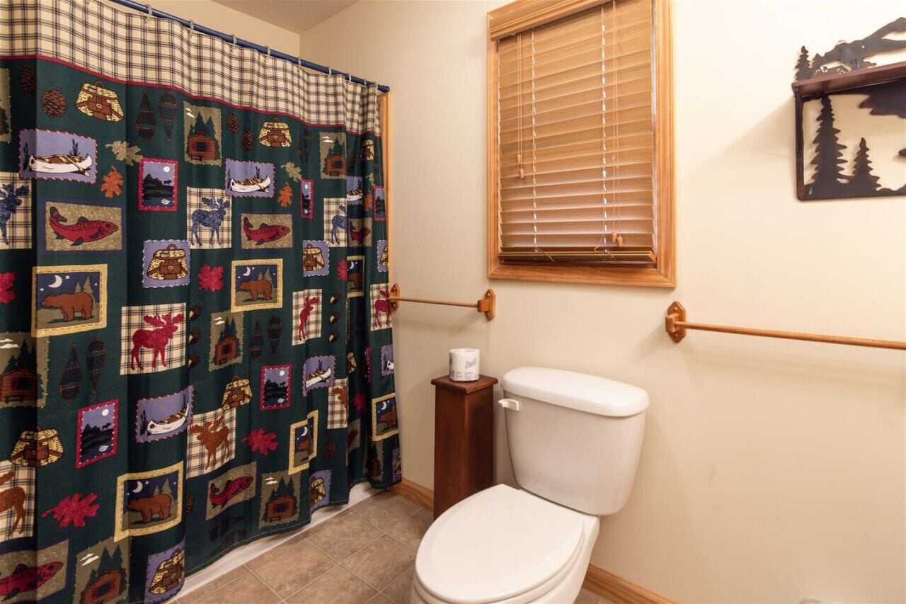 A bathroom with a quilted shower curtain, a toilet, a wooden toilet paper holder, and a window with blinds.