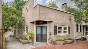 A two-story house with a beige exterior, a black front door, and the number 502 on the door. There are potted plants on the front porch and a small garden bed to the left of the door. - Romantic Cottage in Savannah's Beautiful Historic District! (Savannah)