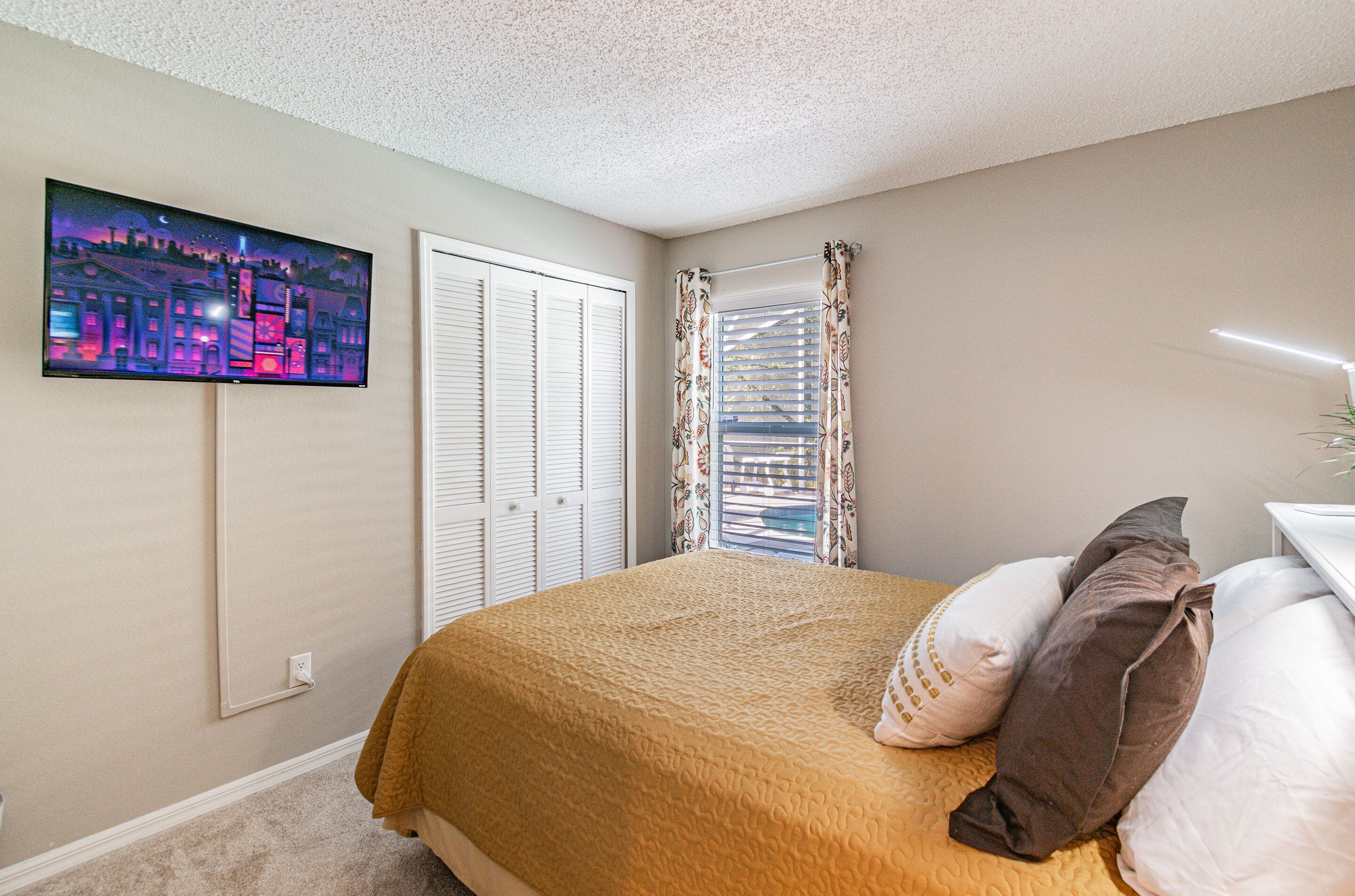 A bedroom with a bed, a television mounted on the wall, a window with curtains, and a door.