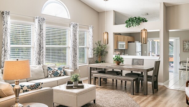 A spacious living room with a sofa, ottoman, and dining table. There is a ceiling fan, large windows with blinds, and a kitchen area visible in the background. - Near Theme Parks! In-Ground 3 BR Pool Home, Sleeps 7, Total Privacy! (Kissimmee)