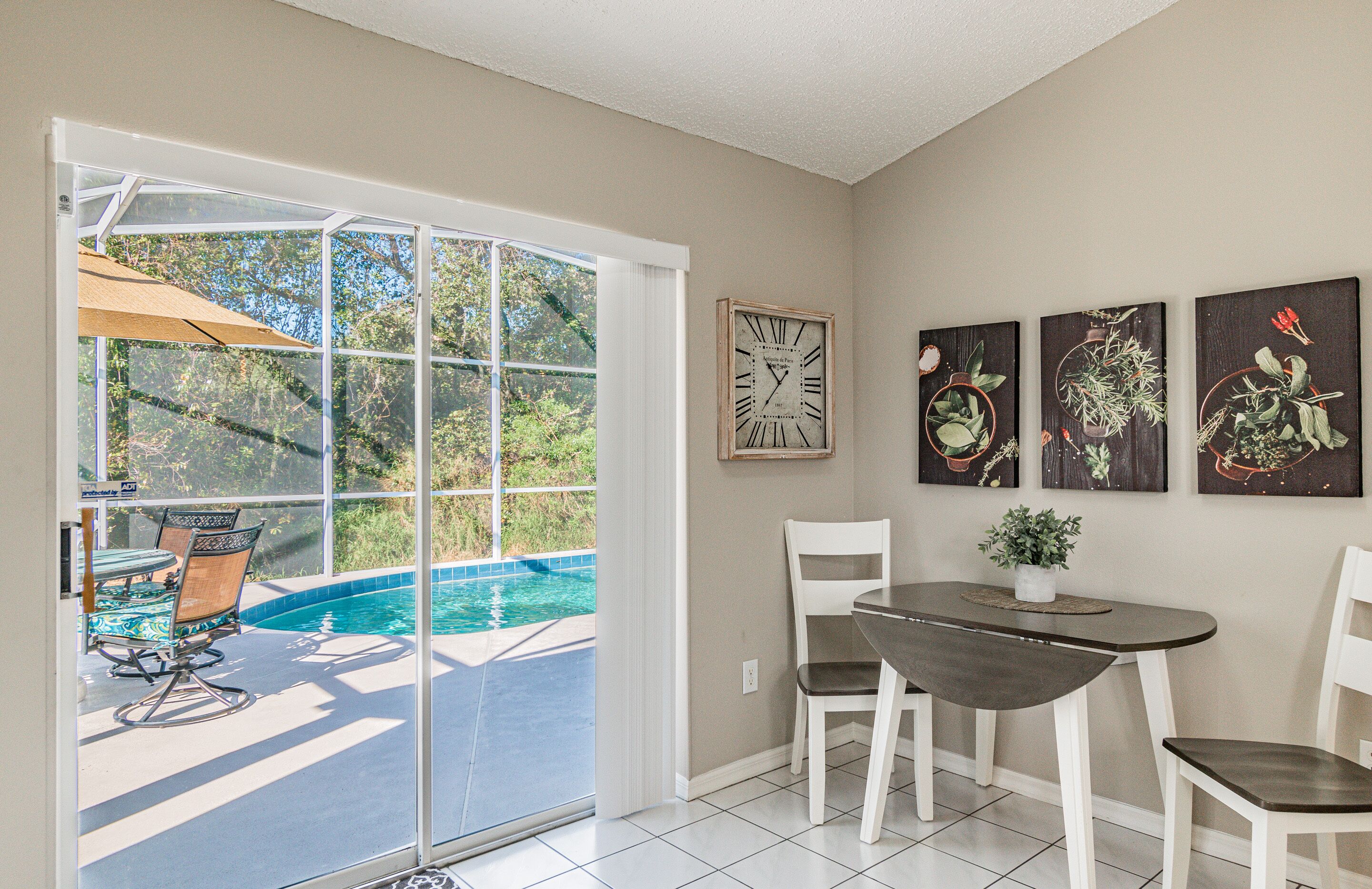 Quarto com porta de vidro deslizante para uma piscina, mesa de jantar com cadeiras e três impressões botânicas emolduradas na parede.