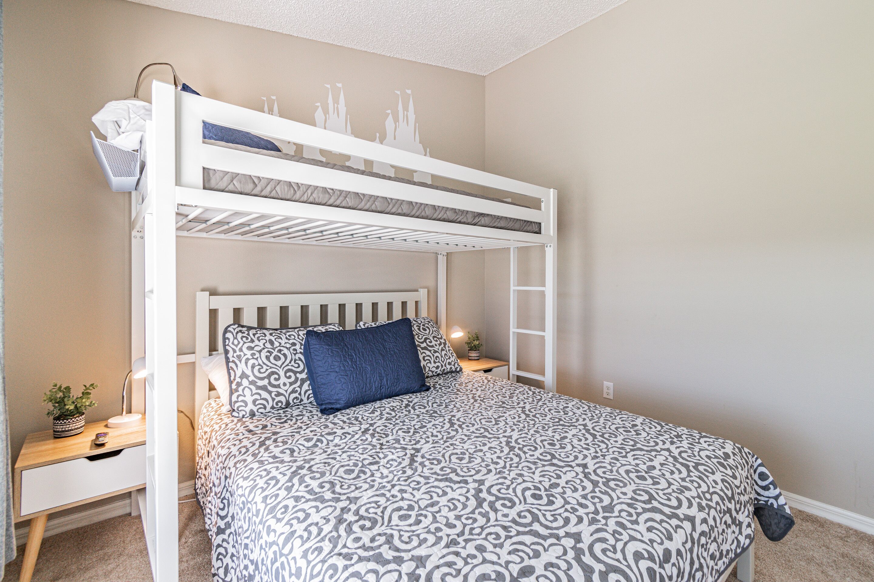 A bunk bed with a patterned bedspread, a bedside table with a lamp, and a small plant.