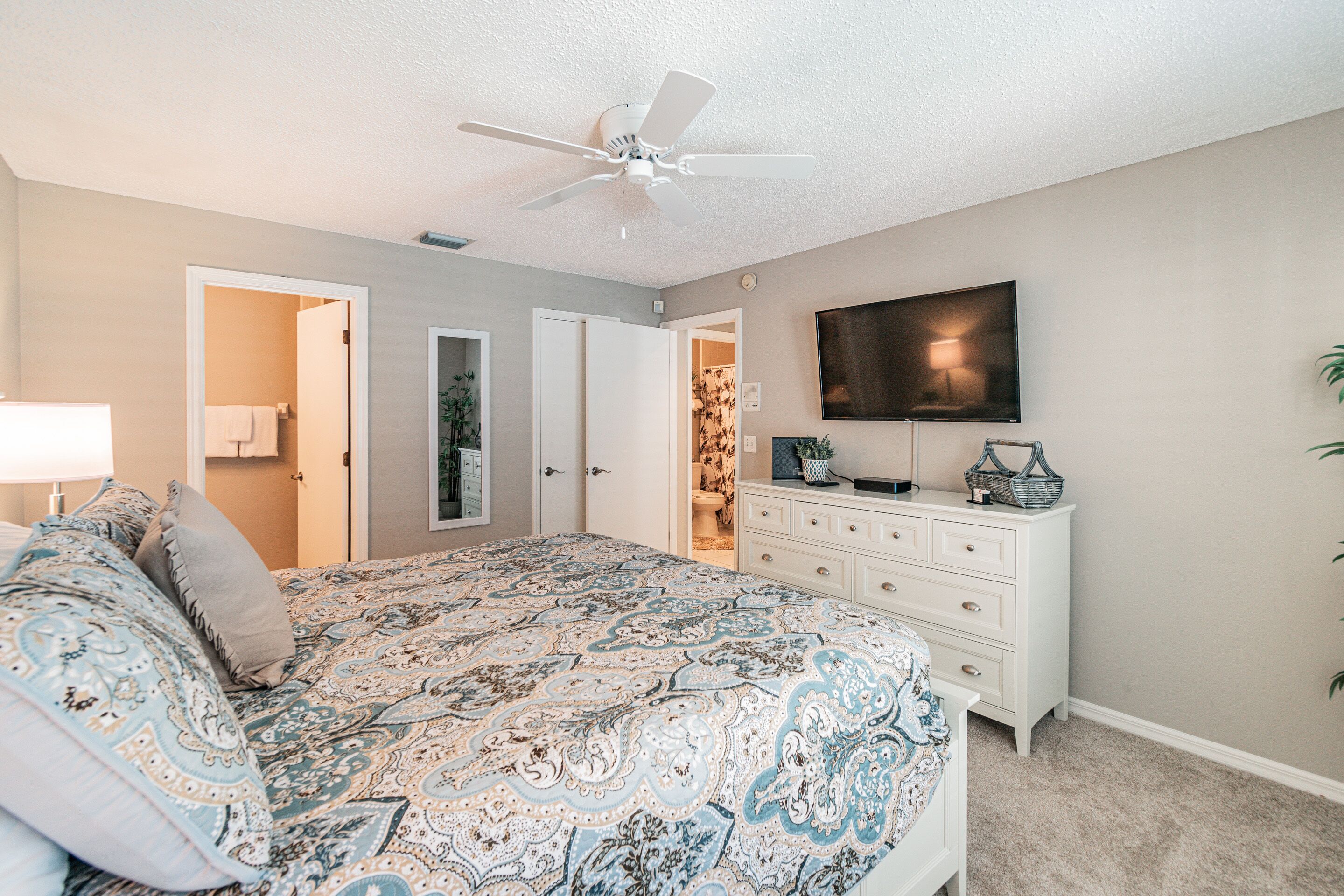 Une chambre à coucher comprenant un lit, une commode, un ventilateur de plafond, un téléviseur et une porte donnant sur une autre pièce.