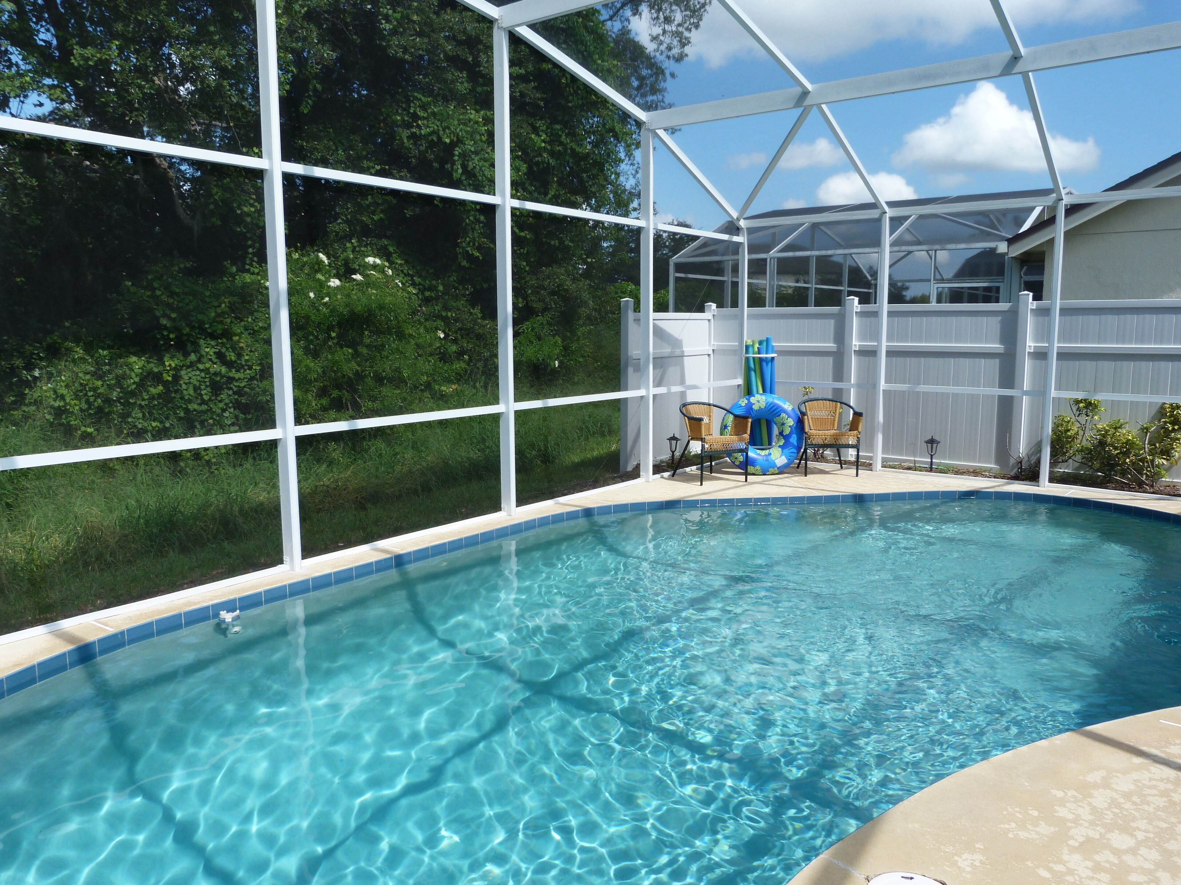 A clear blue pool with a white fence enclosure, two wicker chairs, and a floating ring.